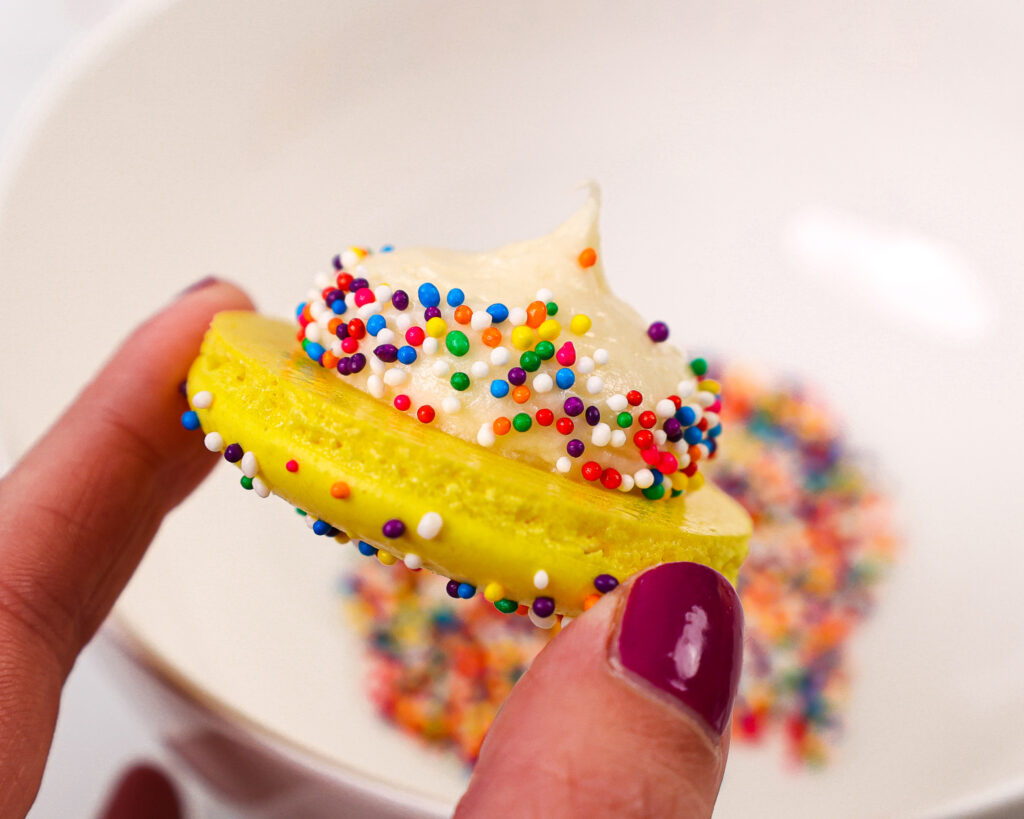 image of a funfetti macaron being filled with cream cheese frosting and rainbow sprinkles