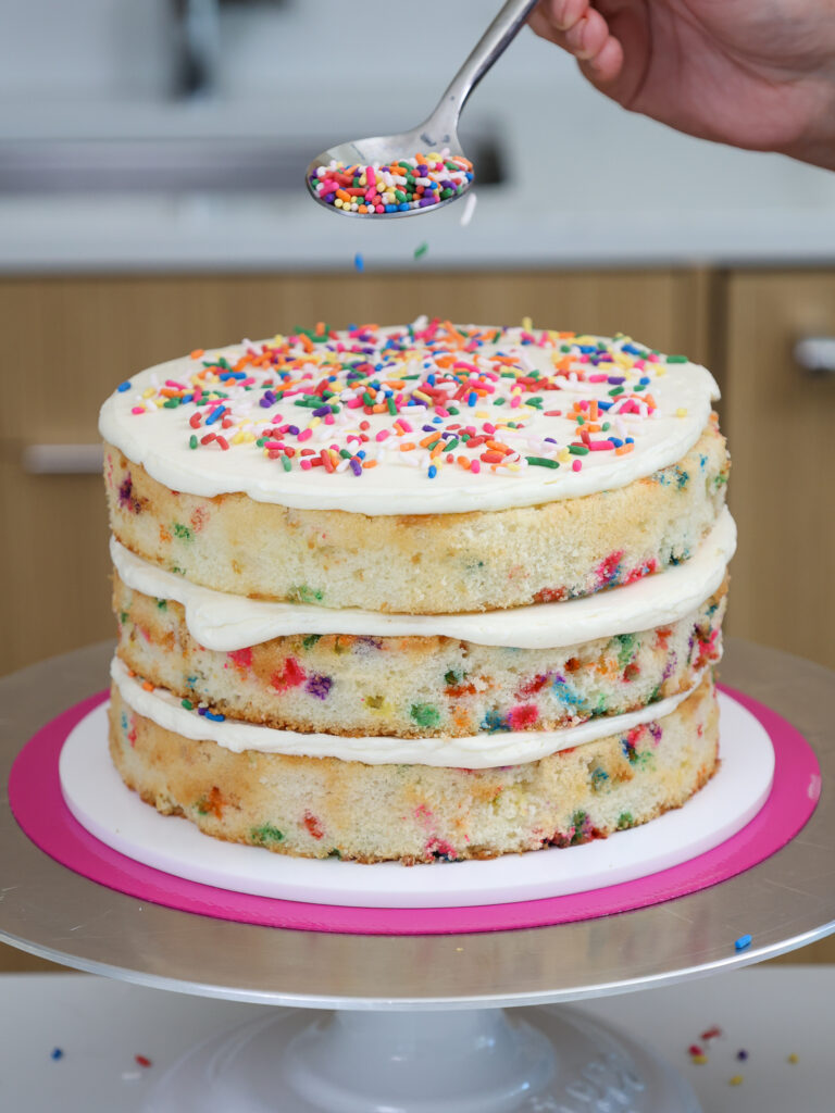 image of a funfetti layer cake being stacked with buttercream frosting and rainbow sprinkles