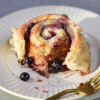 image of a blueberry cinnamon roll on a plate that's been cut into to show how soft and fluffy it is