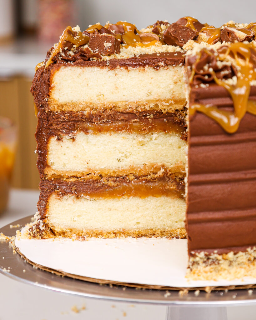 image of a twix cake that's been cut open to show its fluffy vanilla cake layers, thick caramel filling and shortbread crust