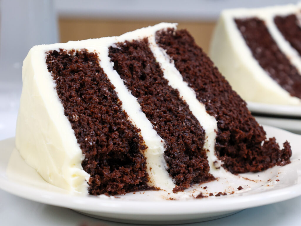 image of a slice of chocolate cake frosted with cream cheese frosting on a plate that's been cut into to show how moist it is