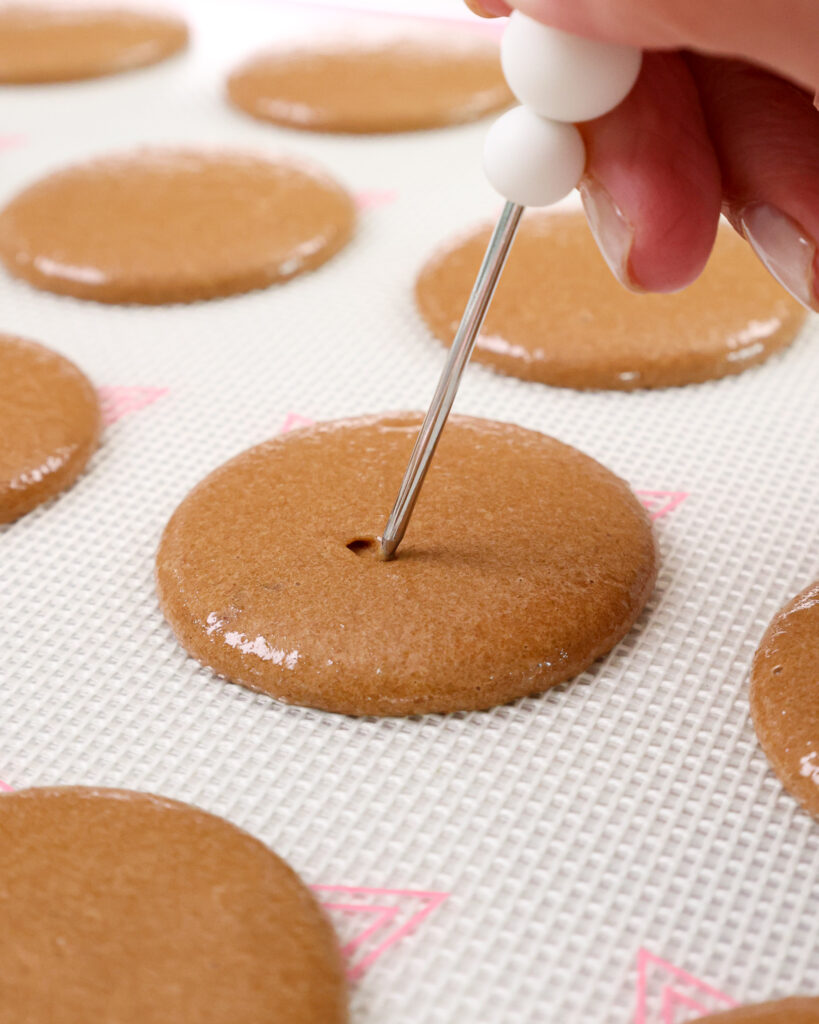 image of a chocolate macaron shell that's having a bubble that was trapped in the shell be popped with a scribe