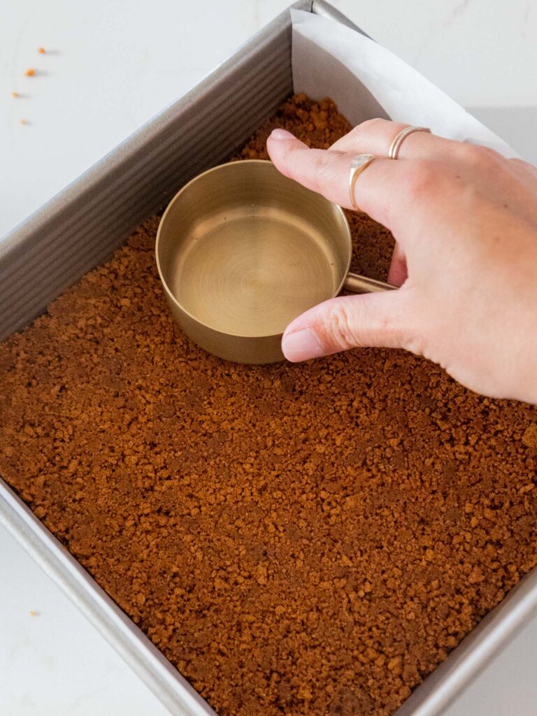image of a biscoff cookie crust being pressed into a square baking pan with the bottom of a measuring cup