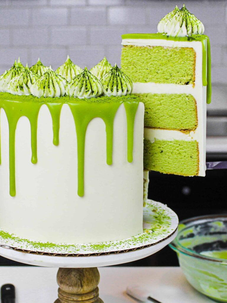 image of matcha cake slice on a plate with pretty matcha drip cake in background