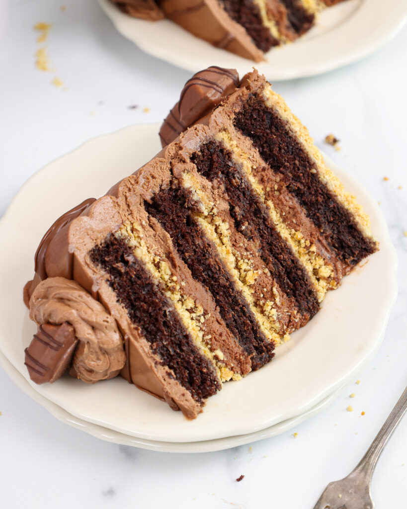 image of a slice of kinder bueno cake that's been cut and put on a plate to show it's crispy shortbread crust, chocolate cake layers and hazelnut mousse filling