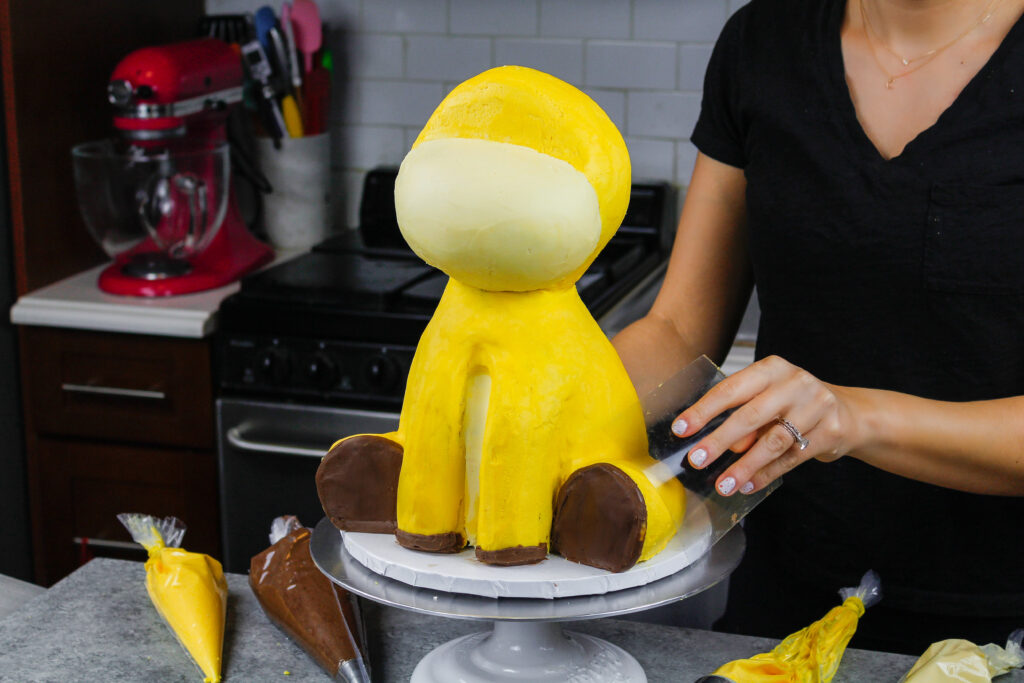 image of yellow buttercream being smoothed onto a giraffe cake using an acetate sheet